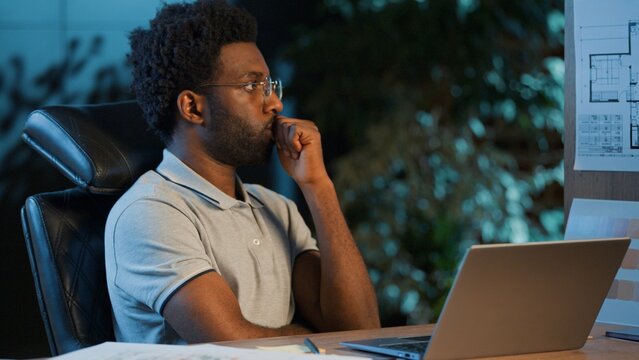 Focused African American business man working laptop thinking at blueprint design project plan consider architecture building scheme at night evening office businessman male guy contractor think idea