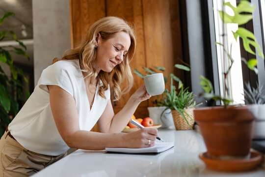 Professional woman taking notes while enjoying a coffee break near a window in a home workspace. Concept of remote work, brainstorming, planning, focus and work-life balance
