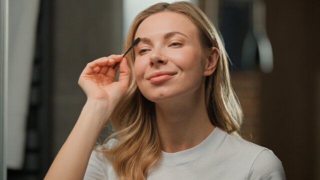 Close up Caucasian adult woman using brush for fixing brushing brows hair make-up beauty product enjoying procedure looking in mirror at home at morning European female combing eyebrows getting ready