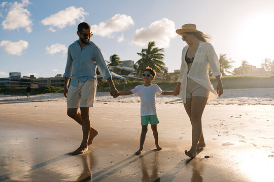 Parents walk hand in hand with their young son along a sunny shoreline, splashing in shallow ocean water and enjoying a relaxed summer family vacation filled with laughter and connection.