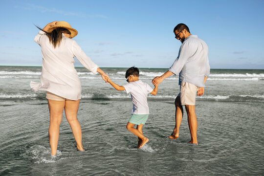 Parents walk hand in hand with their young son along a sunny shoreline, splashing in shallow ocean water and enjoying a relaxed summer family vacation filled with laughter and connection.