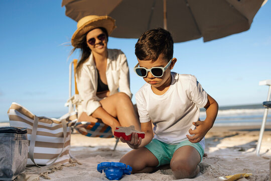 Mother and Son Playing on Sandy Beach Under Umbrella During Sunny Summer Vacation Together