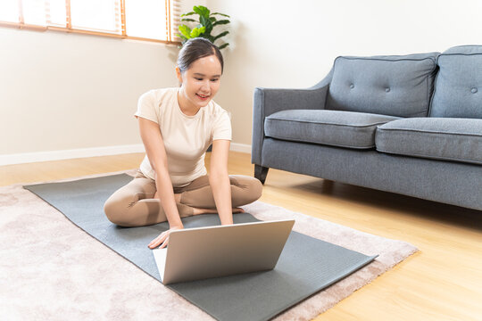 Smiling Asian young woman sitting on a yoga mat and using laptop for online fitness training class at home, digital wellness tutorial and healthy lifestyle in modern living room concept.