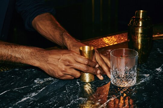 Close up of a mans hands holding a brass jigger on a dark marble bar for cocktail craft and premium mixology concepts