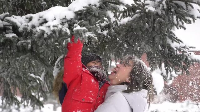 Little Asian boy reaching up to shake snowy pine tree with his mother outdoors.
