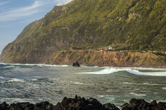 Impresionante vista del mar, olas y acantilado verde con una iglesia y pueblo pintoresco en la costa oeste de la Isla de Flores, Azores