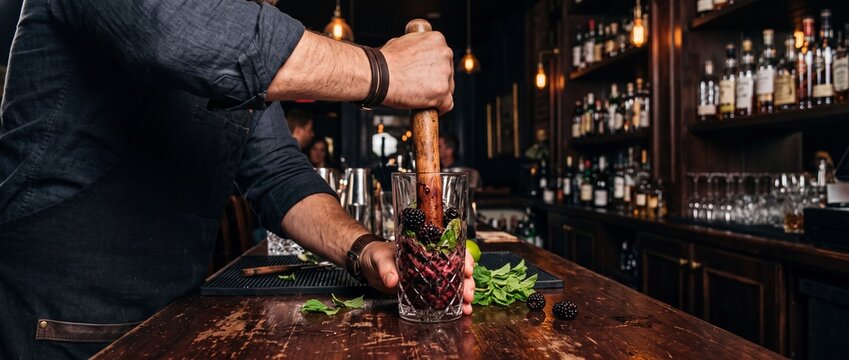 Bartender mashing fresh blackberries with wooden muddler in elegant cut crystal glass to prepare craft cocktail at busy moody bar counter