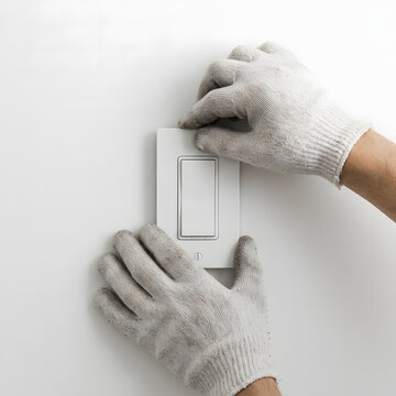 Electrician wearing protective work gloves carefully installs a white plastic wall light switch onto a clean flat interior residential surface