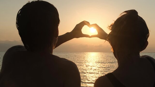 Rear view of a young couple making a heart with their hands against the sunset over the sea horizon. Symbol of true love, devotion, family happiness and romantic relationship.