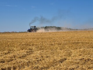 Naklejka premium Blue tractor working in harvested wheat stubble field with dust cloud and smoke