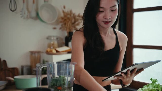 Young athletic woman adding fresh vegetables and greens into a blender while consulting a recipe on a digital tablet in a modern kitchen