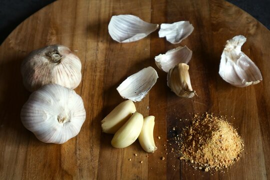 Two whole garlic cloves (Allium sativum), several peeled garlic cloves along with their skins, and a mound of garlic powder on a wooden cutting board.