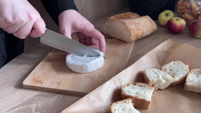 Cutting round brie cheese into wedges on wooden cutting board during homemade food preparation in home kitchen, slicing soft cheese with kitchen knife using human hands