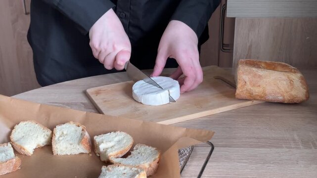 Cutting round brie cheese into wedges with kitchen knife on wooden cutting board during homemade food preparation in modern home kitchen, close-up cooking process with human hands preparing soft chees