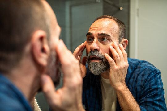 Concerned middle-aged man examining face in bathroom mirror