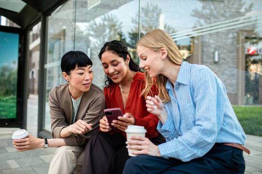 Three women sharing smartphone and coffee at bus stop