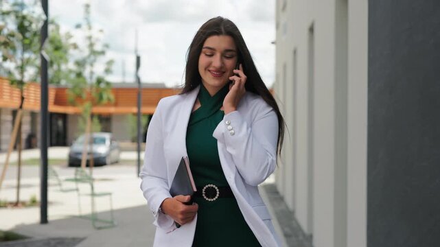 Beautiful young business woman walking towards camera and talking on phone outdoors
