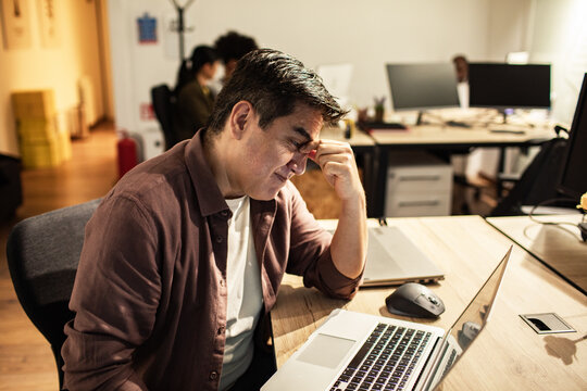 Stressed man rubbing eyes at laptop in modern office
