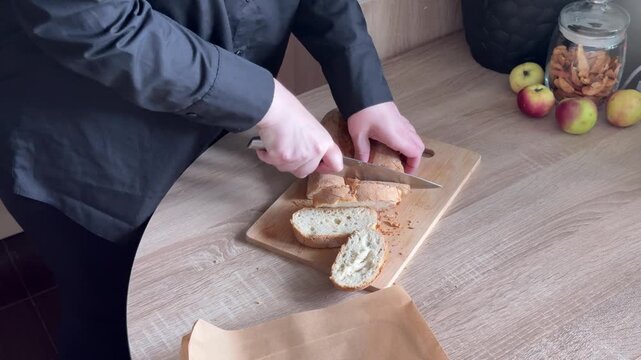Slicing fresh loaf of bread on wooden cutting board with kitchen knife during homemade food preparation in home kitchen, close-up cooking process with human hands