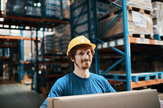 Smiling warehouse worker with hard hat carrying box in aisle