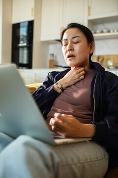 Woman holding neck while using laptop in home kitchen