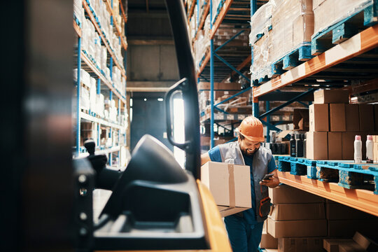 Warehouse worker carrying box and checking smartphone in aisle