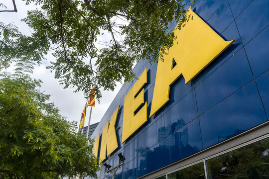 Logotipo amarillo de IKEA sobre la fachada azul de una tienda IKEA, enmarcado por &aacute;rboles verdes y fotografiado desde un &aacute;ngulo bajo bajo un cielo nublado en Barcelona, Espa&ntilde;a.