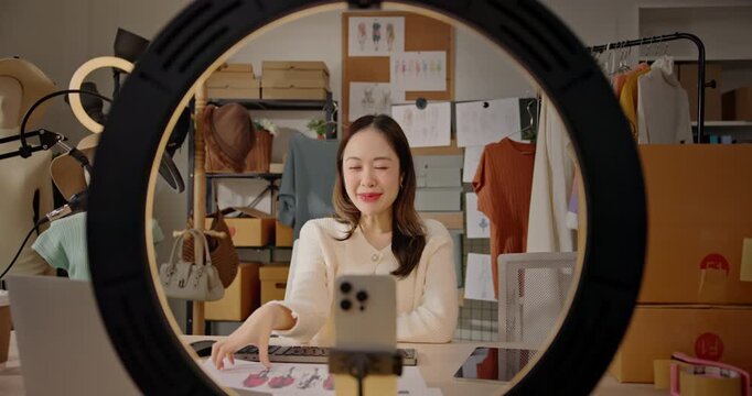 A stylish Asian woman fashion designer, influencer livestreams from studio workspace, Promotes her brand and communicates with online customers on social media platform. entrepreneurship, e-commerce.