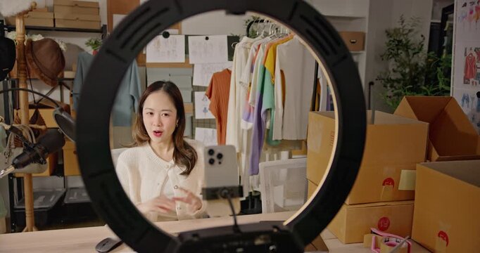 A stylish Asian woman fashion designer, influencer livestreams from studio workspace, Promotes her brand and communicates with online customers on social media platform. entrepreneurship, e-commerce.