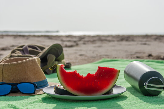 Bitten watermelon on plate with hat sunglasses and water bottle on beach