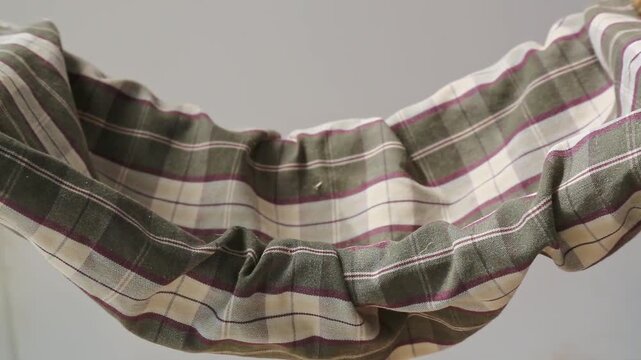 A small grayish brown mouse stands on a plaid fabric hammock, climbing upward and looking around alertly, then moving toward the edge and shaking its fur briefly indoors.