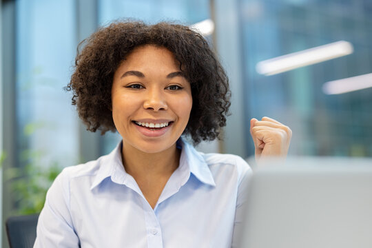 Happy young African American woman sitting in the office in front of a laptop and rejoicing at the news she read, showing a victory gesture with her hand