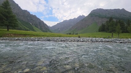 Dramatic mountain valley river, clear flowing water over stones, green alpine meadow and peaks under cloudy sky © sumblistan