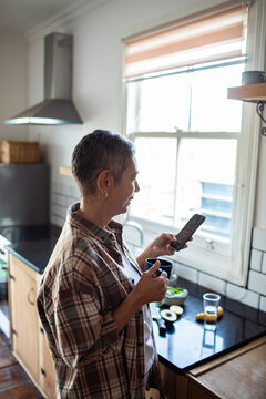 Mature woman using smartphone and drinking coffee in home kitchen