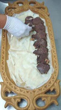 Chef in white gloves arranging sliced horsemeat sausage kazi and boiled beef over dough sheets for traditional uzbek naryn beshbarmak dish on a carved wooden platter, vertical video.