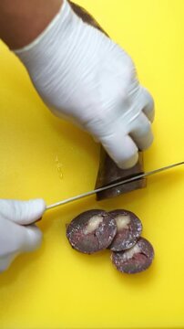Top view of professional chef in white gloves slicing traditional uzbek kazi horsemeat sausage on a bright yellow cutting board, vertical video.