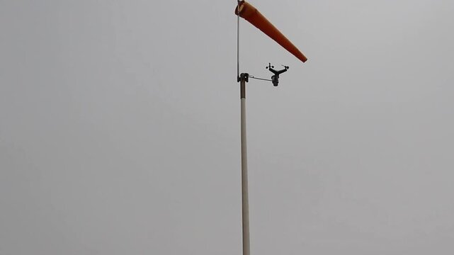 Orange Windsock And Cup Anemometer Spinning On Top Of Pole Against Overcast Sky
