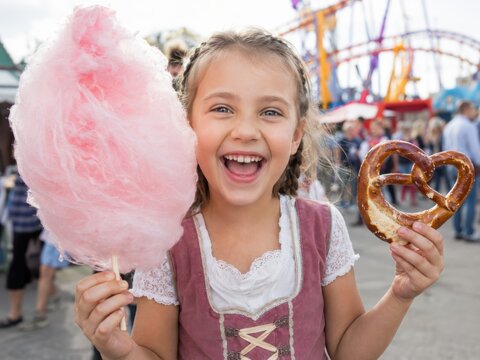 Lachendes M&auml;dchen im Dirndl h&auml;lt riesige rosa Zuckerwatte und eine herzf&ouml;rmige Brezel vor bunten Fahrgesch&auml;ften &ndash; fr&ouml;hlicher Familienmoment auf dem Volksfest.