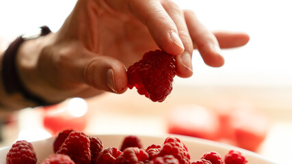 Human hand holding a fresh red raspberry on an open palm over a white bowl filled with ripe berries. © kyrylovasyliev