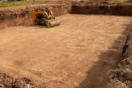 Skid steer loader with roller attachment compacting ground base in deep excavation pit