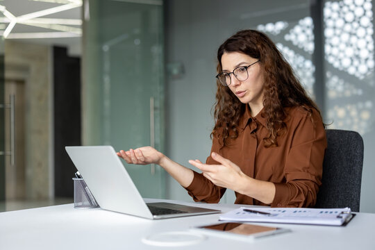 Young businesswoman with glasses having a serious business video conference on a laptop, gesturing with her hands while explaining or discussing an important topic during remote work
