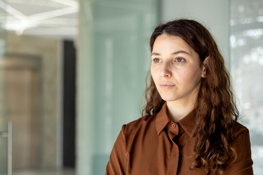 Woman with long brown hair wearing a shirt, looking away thoughtfully while standing in a bright corporate office interior, representing focus, success, and forward-thinking