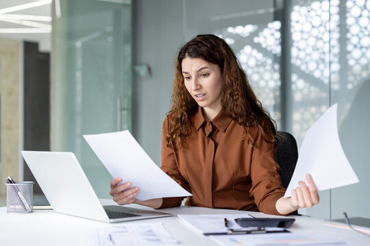 Young woman feeling overwhelmed and frustrated while working with business documents and a laptop, experiencing stress and confusion in an office setting