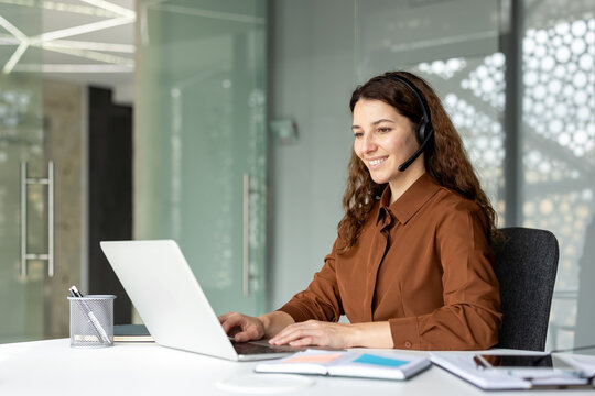 Young woman smiling and communicating with clients using a laptop and headset, working remotely from a modern office environment and providing effective customer support