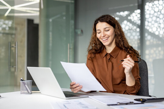 Young businesswoman with curly hair smiling while reviewing documents and working on a laptop at her office desk, representing productivity and professional success