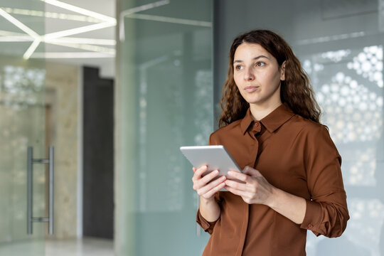 Young woman standing in a modern office environment, holding a digital tablet and looking away thoughtfully, embodying technology integration and business planning