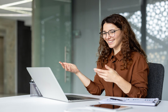 Young woman with curly hair and glasses smiling and gesturing during a video call on her laptop, communicating while working remotely from a modern office