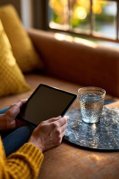 Hands holding blank tablet near sofa and glass of water in cozy home setting