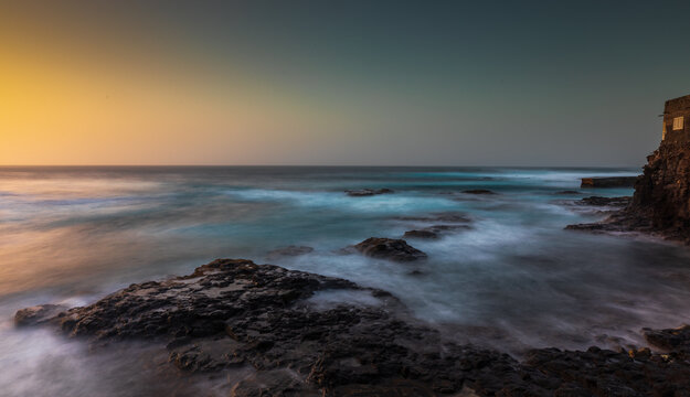 View of the rocky coastline of Santo Antao at sunset with crashing waves and a cliffside building in Ponta do Sol, Ribeira Grande, Cabo Verde.