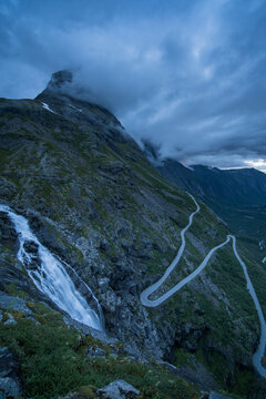 View of the dramatic hairpin bends of the Trollstigen mountain road and Stigfossen waterfall cascading down rugged cliffs under a cloudy sky in Andalsnes, More og Romsdal, Norway.